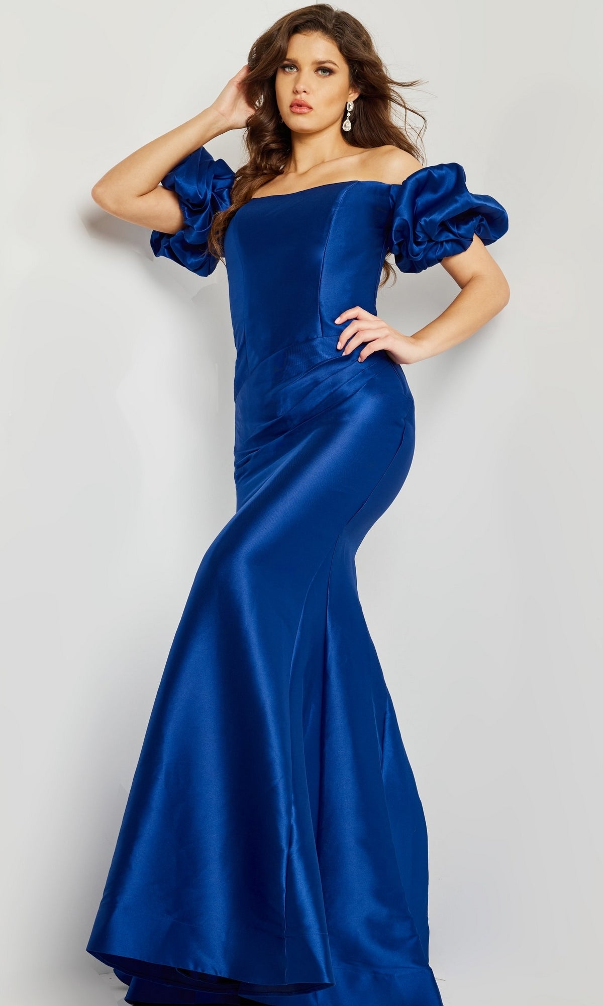 A woman models the Jovani 08361 Puff-Sleeve Mikado Evening Dress, a floor-length royal blue gown with off-the-shoulder puffed sleeves and a mermaid silhouette, set against a neutral background.